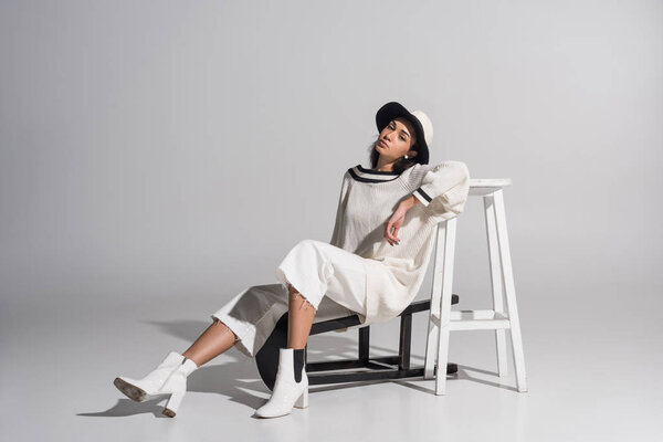beautiful african american girl in fashionable white clothes and hat sitting on black and white chairs on white