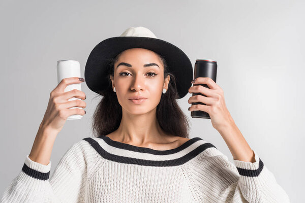 attractive african american girl in stylish white sweater and hat holding white and black cans and looking at camera isolated on white