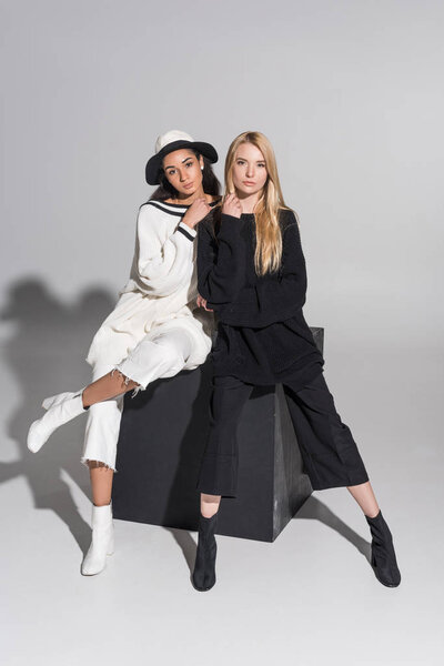 beautiful multicultural friends in black and white clothes sitting on black cube and looking at camera on white