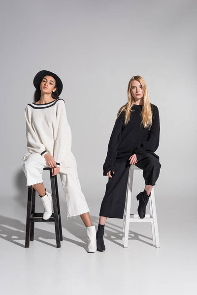 attractive multiethnic friends in black and white clothes sitting on chairs and looking at camera on white