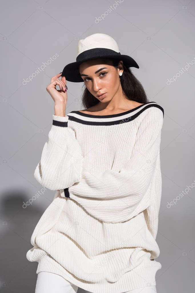 attractive african american girl in trendy white clothes touching hat and looking at camera on white
