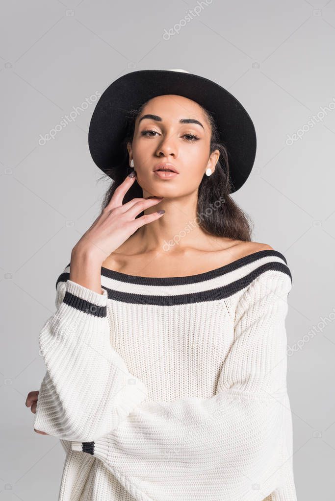 attractive african american girl in stylish white sweater and hat touching face and looking at camera isolated on white