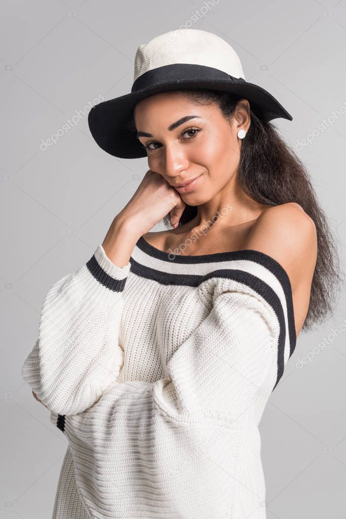 attractive cheerful african american girl in stylish white sweater and hat looking at camera isolated on white