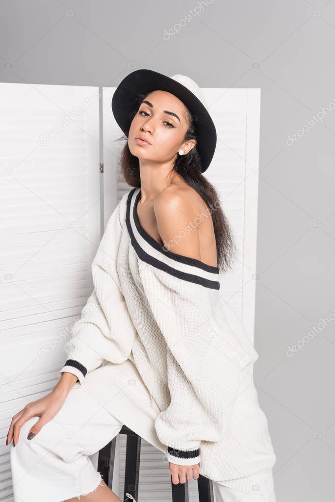 beautiful african american girl in stylish white clothes and hat sitting and posing near folding screen on white