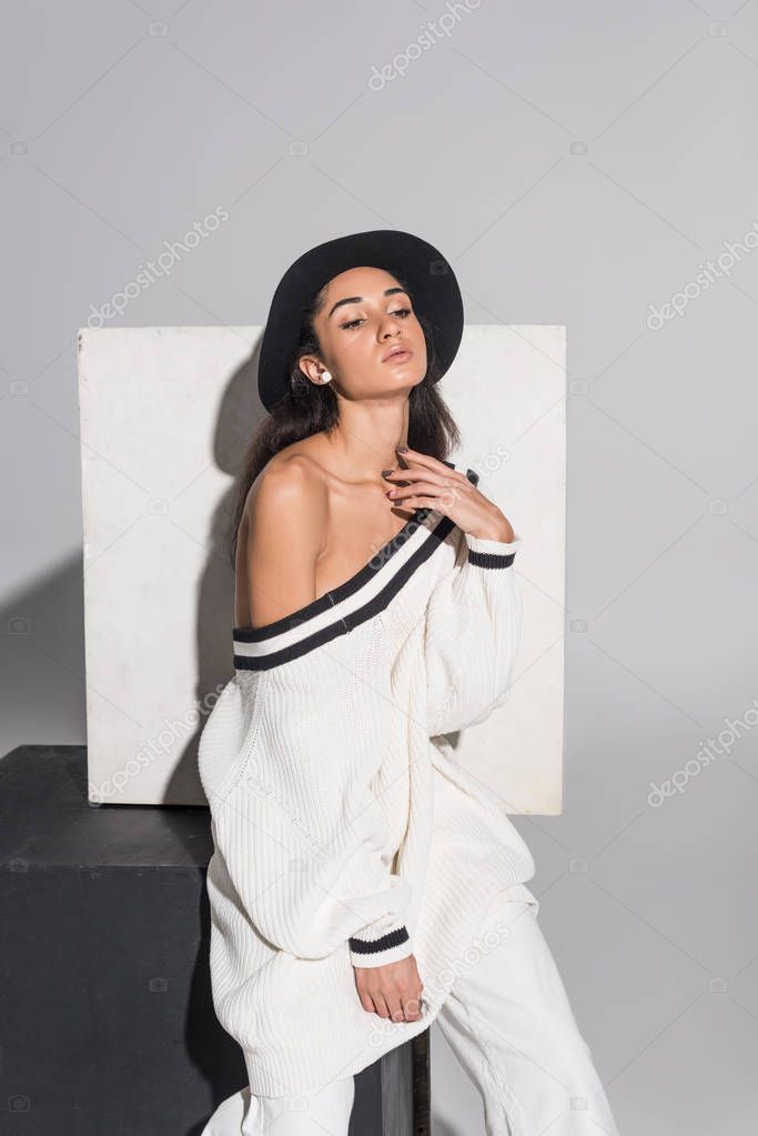 attractive african american girl in stylish white clothes and hat sitting on black cube and looking away on white