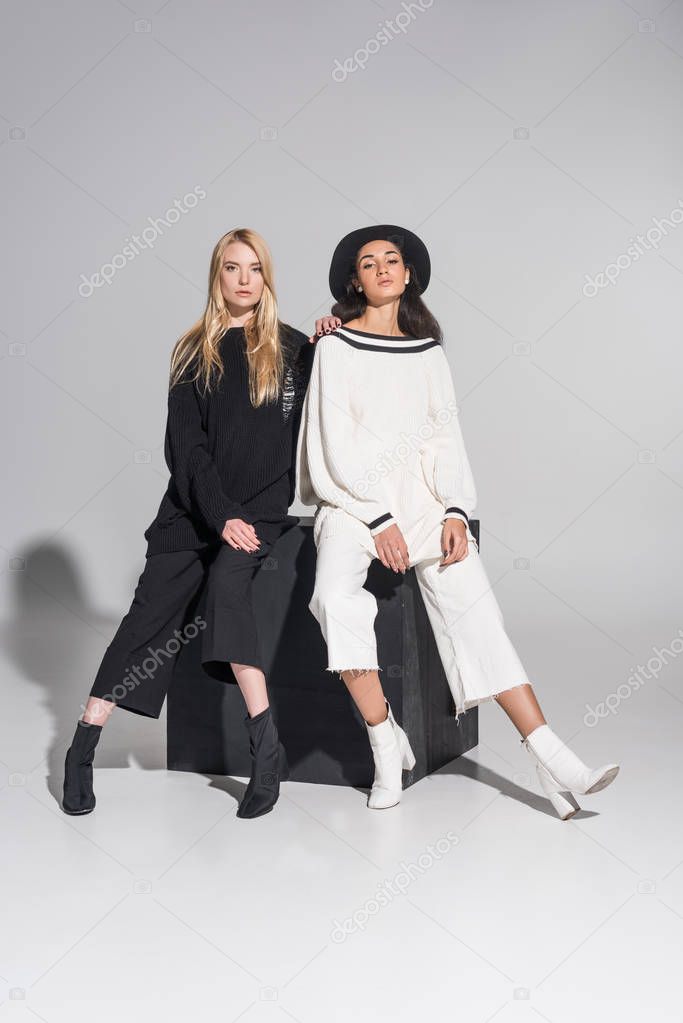 beautiful multiethnic women in black and white clothes sitting on black cube and looking at camera on white