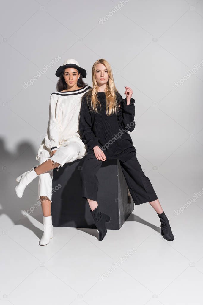 beautiful multicultural female models in fashionable black and white clothes sitting on black cube and looking at camera on white