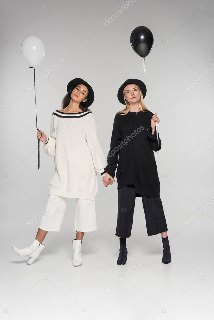 beautiful multicultural lesbian couple in black and white clothes holding hands and two balloons on white, looking at camera