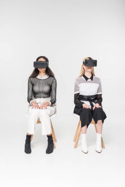 multicultural women in virtual reality headsets sitting on chairs isolated on white clipart