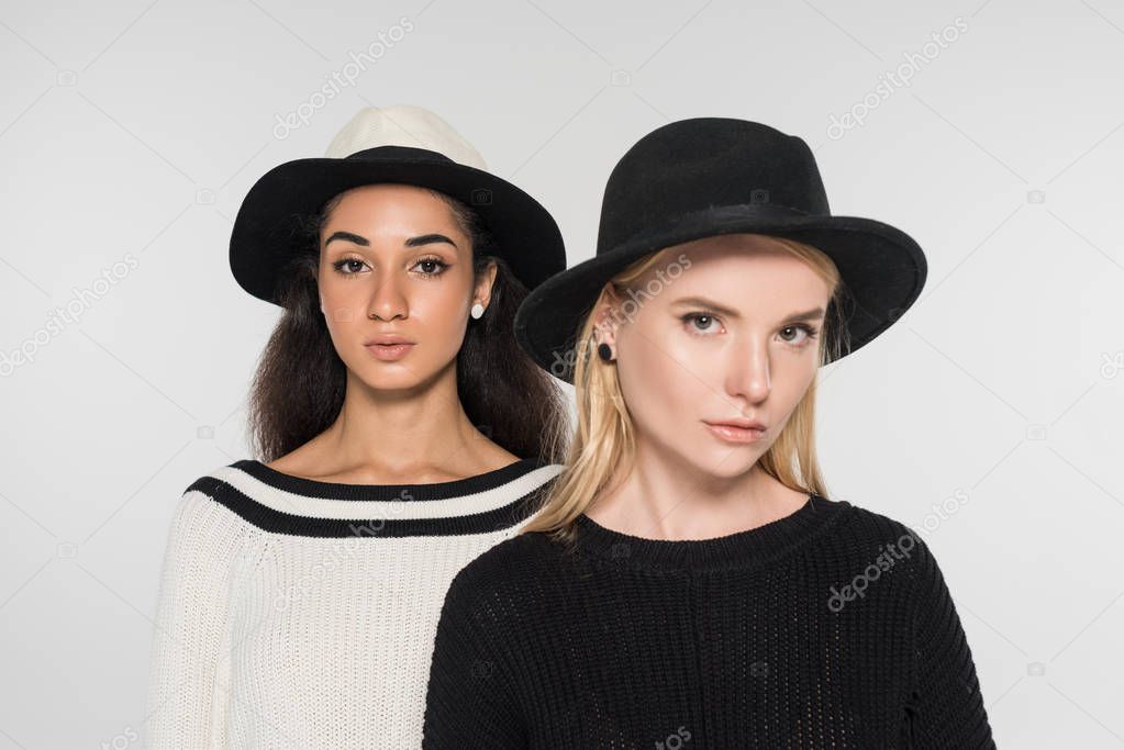 portrait of beautiful multiethnic women in black and white clothes and hats looking at camera isolated on white