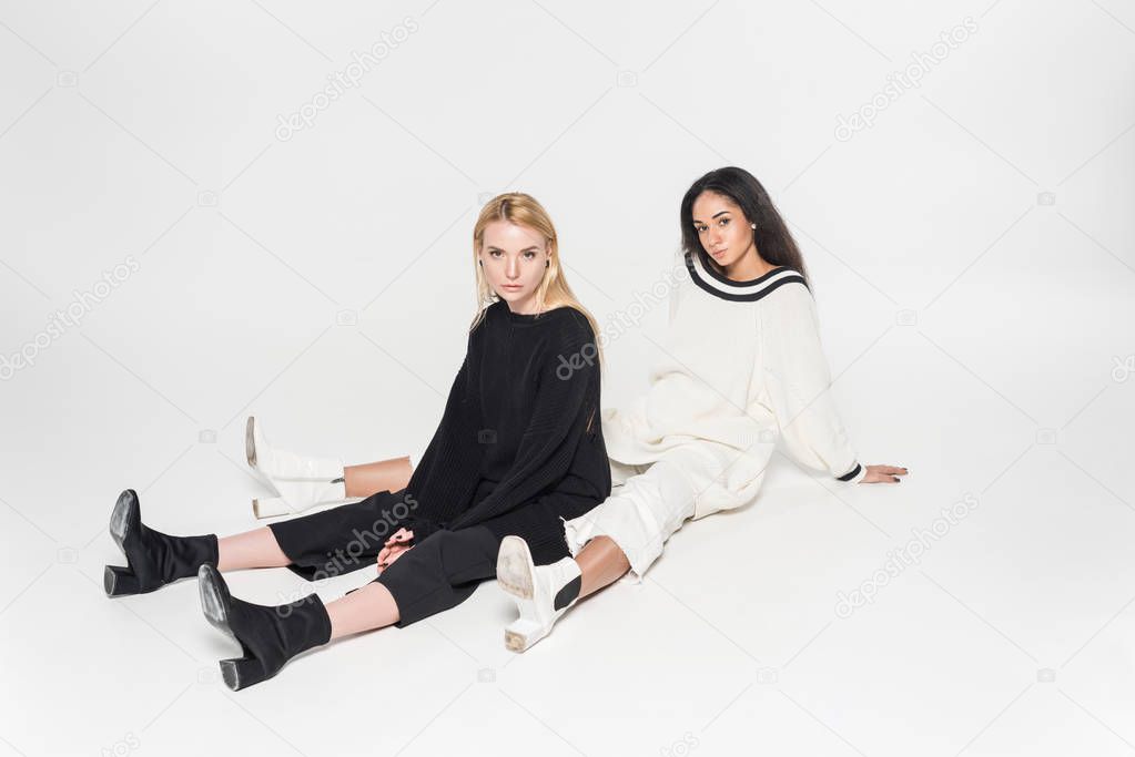 attractive multicultural friends in black and white clothes sitting and looking at camera on white