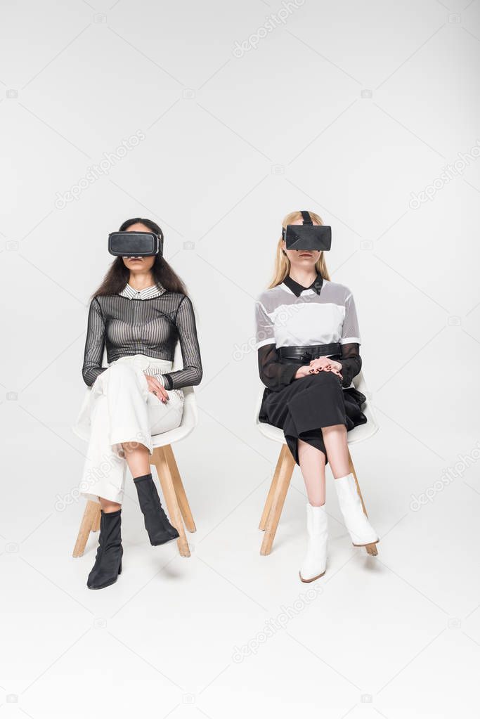 attractive multiethnic women in virtual reality headsets sitting on chairs isolated on white