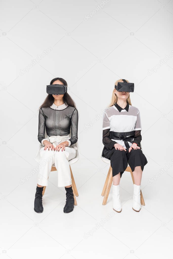multicultural women in virtual reality headsets sitting on chairs isolated on white