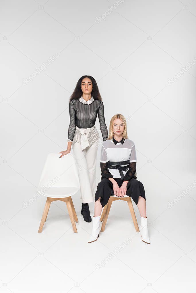 attractive multiethnic women in black and white clothes sitting on chairs and looking at camera isolated on white