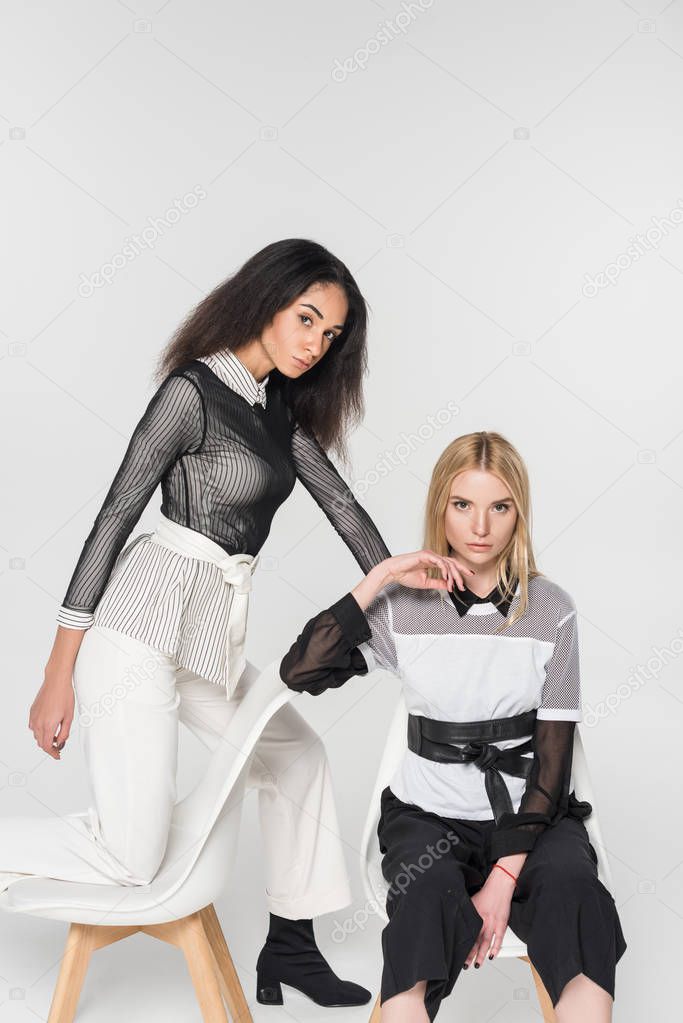 attractive multiethnic women in black and white clothes posing on chairs isolated on white