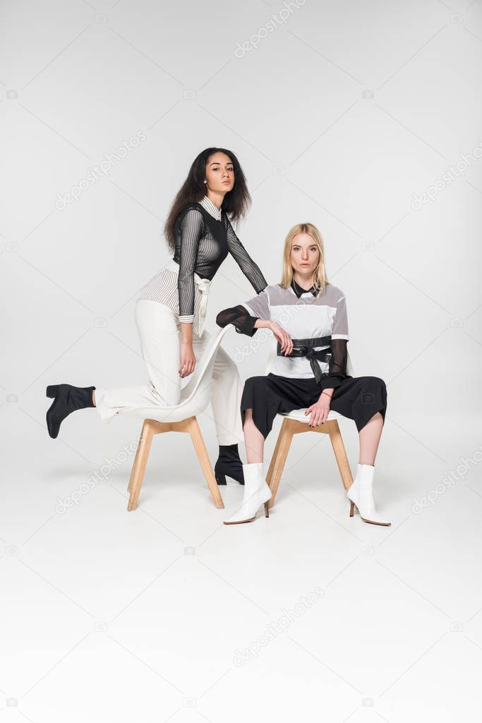attractive multiethnic women in stylish black and white clothes sitting on chairs isolated on white