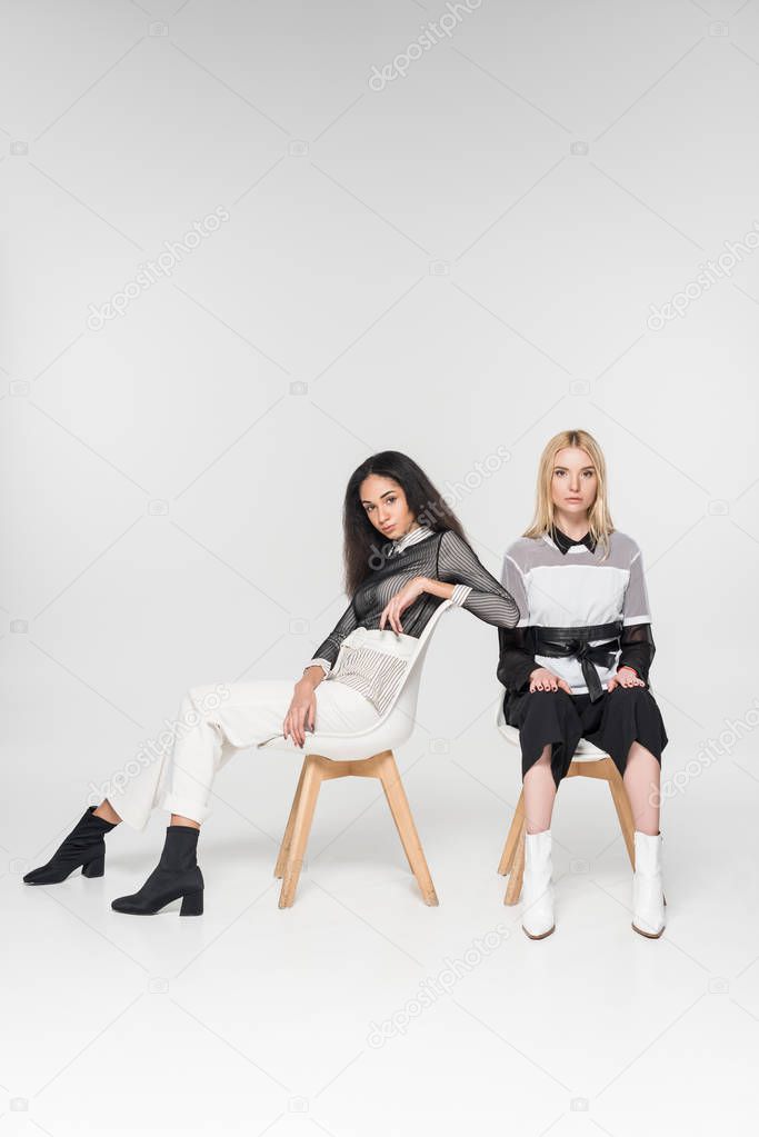 attractive multiethnic friends in black and white clothes sitting on chairs and looking at camera isolated on white