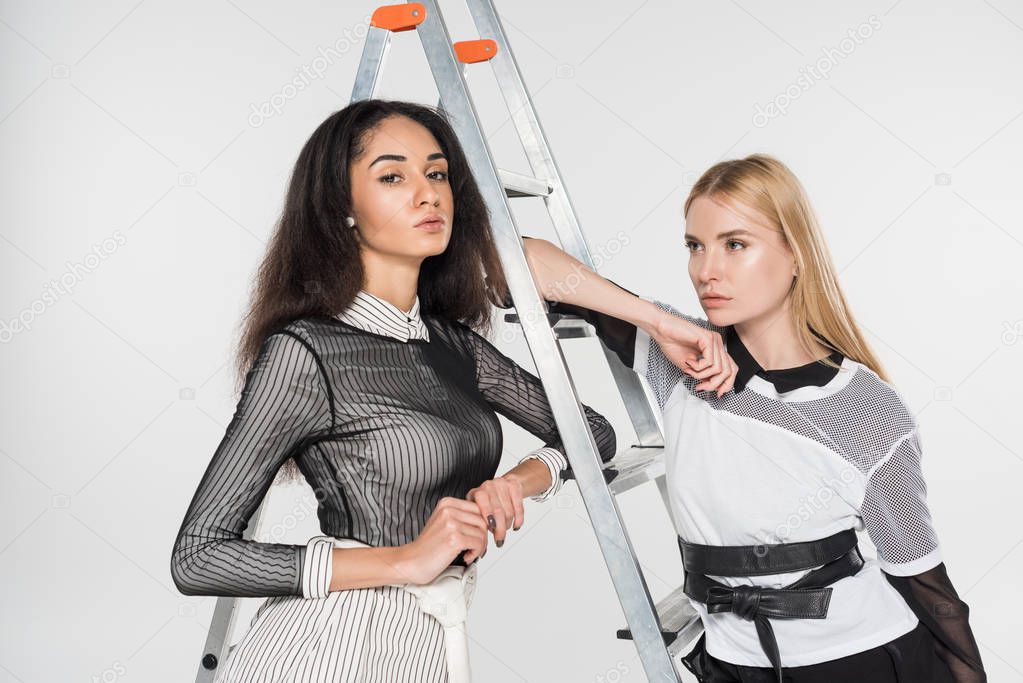 attractive multicultural girls in black and white clothes leaning on ladder isolated on white