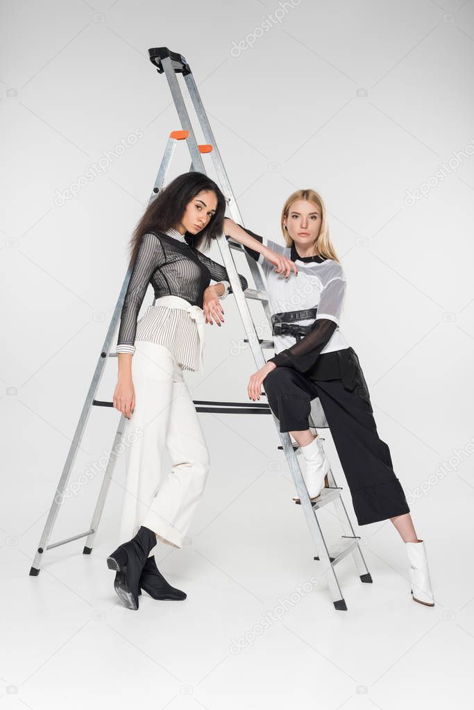 attractive multiethnic female models in black and white clothes leaning on ladder and looking at camera isolated on white