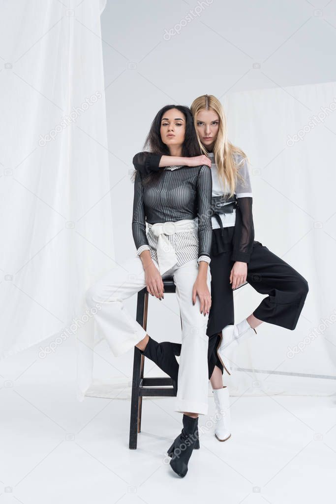 attractive interracial girlfriends in black and white clothes sitting on chairs and hugging on white