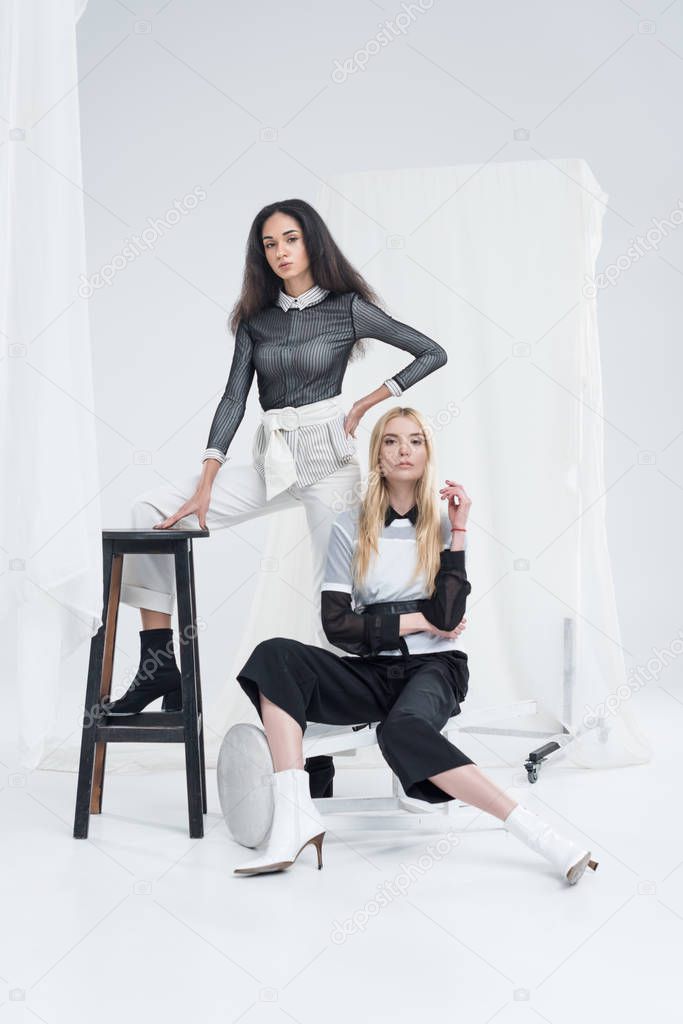 attractive multiethnic women in stylish black and white clothes posing on chairs near tulle on white