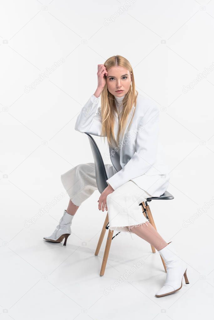attractive caucasian blonde woman in fashionable white clothes sitting on chair and looking at camera isolated on white