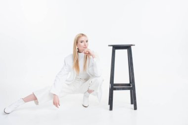 attractive caucasian blonde woman in fashionable white clothes squatting near black chair isolated on white clipart