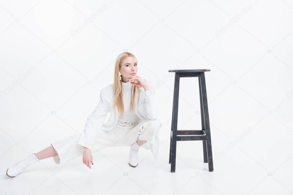 attractive caucasian blonde woman in fashionable white clothes squatting near black chair isolated on white