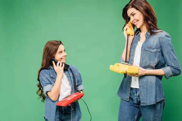beautiful mother and daughter talking on vintage telephones isolated on green