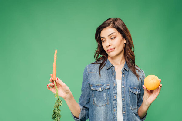 skeptical woman in denim choosing between orange and carrot isolated on green