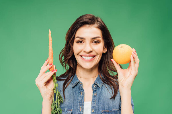 smiling woman in denim holding orange and carrot isolated on green