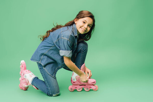 cute child putting on rollerblades and looking at camera on green background