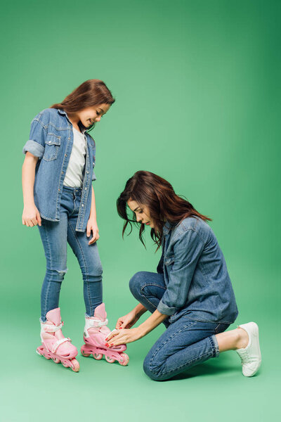 mother in denim putting roller blades on daughter on green background
