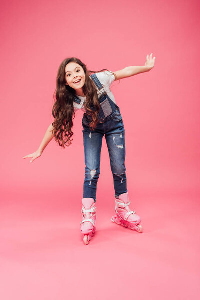 cute smiling child in overalls rollerblading with outstretched hands isolated on pink background