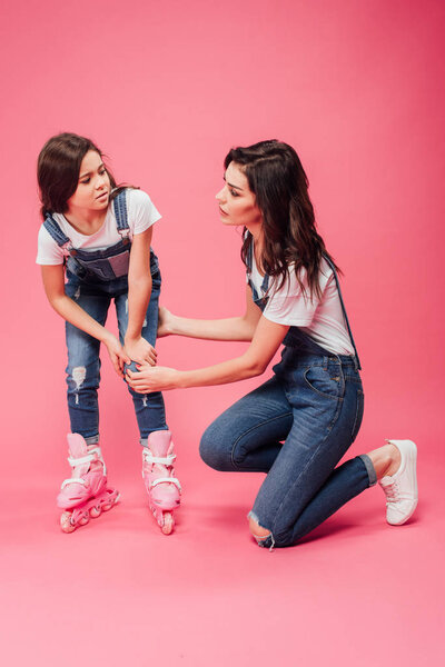 mother checking on hurt knee of daughter in roller blades on pink background