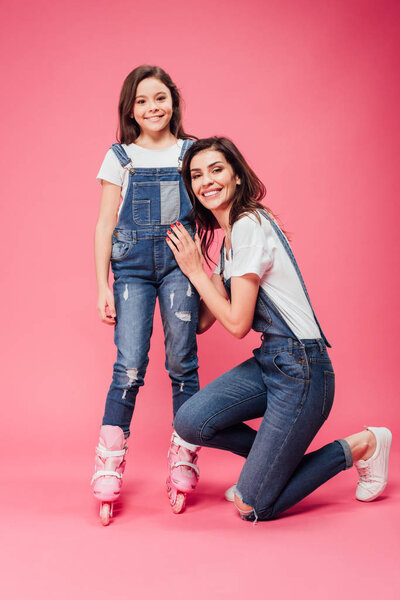 happy mother hugging daughter in roller blades and looking at camera on pink background