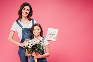 happy mother and daughter smiling while holding flowers and greeting card on mothers day isolated on pink  clipart
