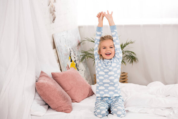 smiling adorable kid in pajamas sitting on bed with hands in air 