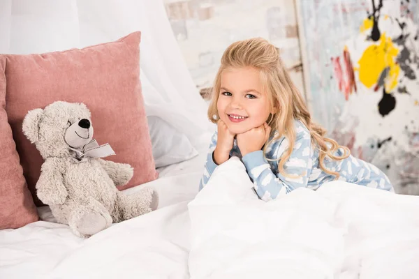 可爱的孩子与泰迪熊在床上看着相机 — 图库照片
