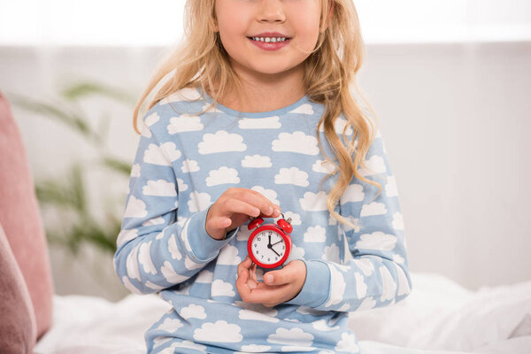 partial view of adorable kid sitting on bed with small clock in hands
