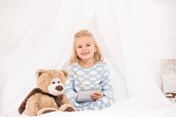 可爱的孩子在睡衣坐在床上与笔记本和泰迪熊 — 图库照片