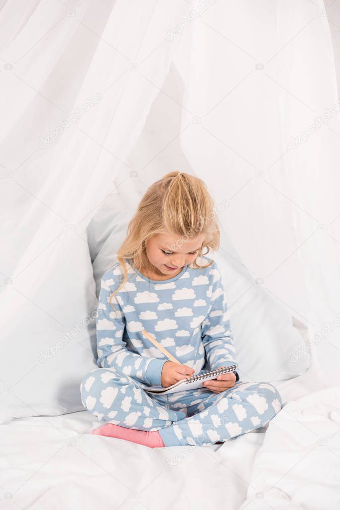 cute kid in pajamas sitting on bed with crossed legs and writing in notebook