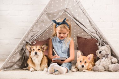 Kızılderili çadırı corgi köpek ve not defterinde yazma ile oturma sevimli çocuk 