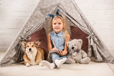 Kızılderili çadırı corgi köpek ve not defterinde yazma ile oturma sevimli çocuk