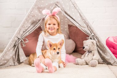 cute child in bunny ears headband sitting with corgi dog and teddy bear in wigwam clipart