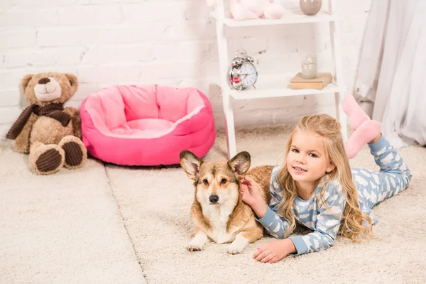 Улыбающийся Ребенок Пижаме Смотрит Камеру Гладит Валлийскую Собаку Корги Дома — Бесплатное стоковое фото