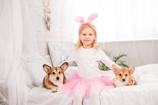 улыбающийся ребенок в кроличьих ушах повязка сидя с валлийскими корги собак на кровати дома
 