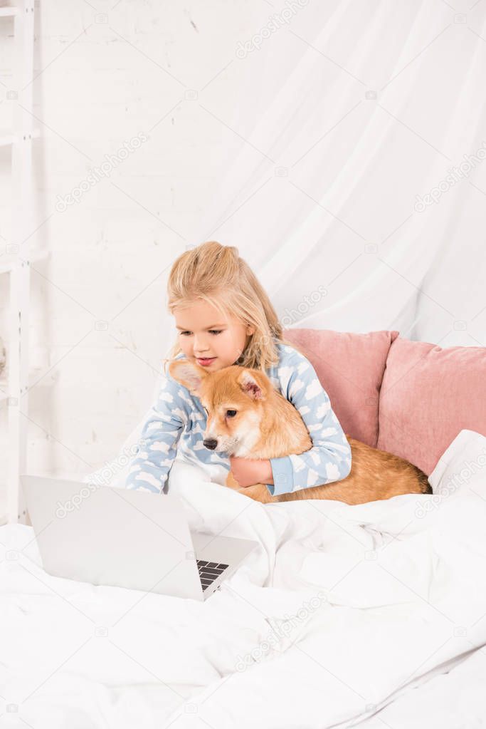 adorable child lying in bed, hugging corgi dog and using laptop at home