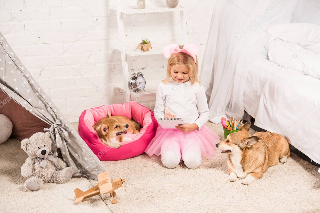 child in bunny ears headband sitting with welsh corgi dogs at home and writing in notebook