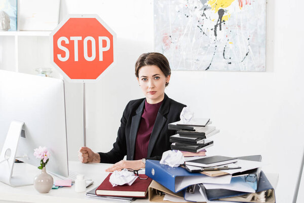 attractive businesswoman sitting at table and holding stop sign in office